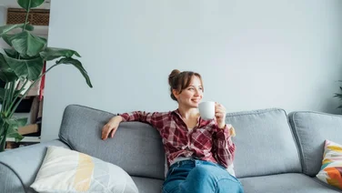 Young smiling woman sitting on sofa against a wall and looking away while holding a mug