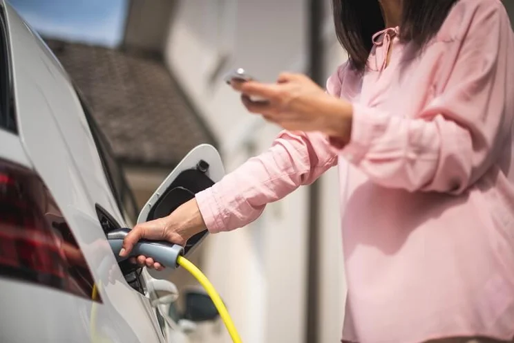 A woman wearing a pink shirt charging her electric car at home, while checking her phone