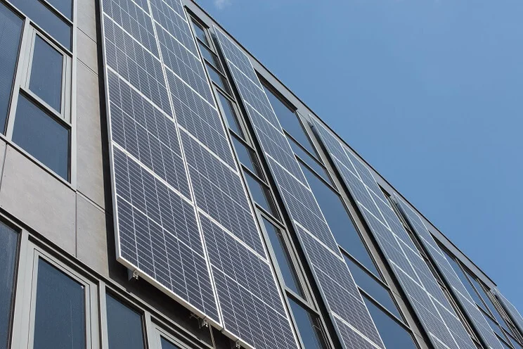 Lots of blue solar panels lined up vertically on the side of a building with windows on it