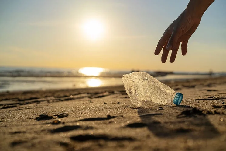 Close up of man's hand picking up crumpled plastic bottle on beach at sunset