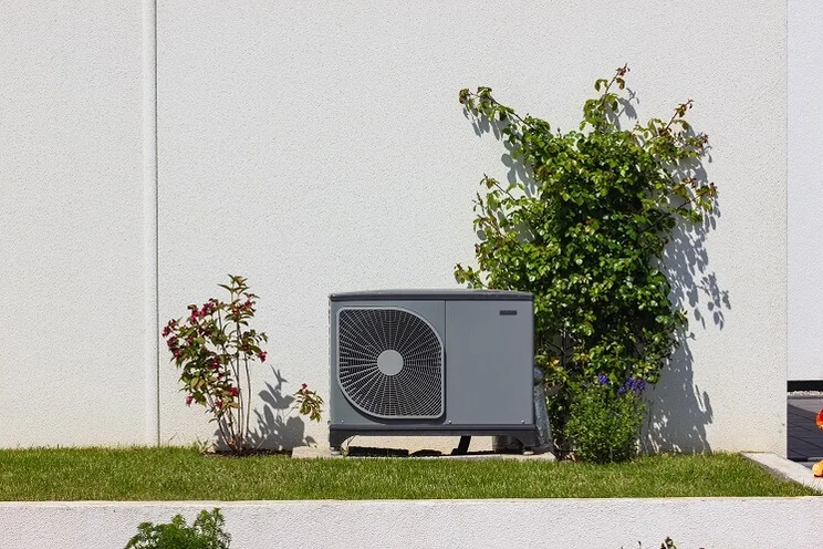 air source heat pump unit outside house with white wall, grass lawn, and shrubs
