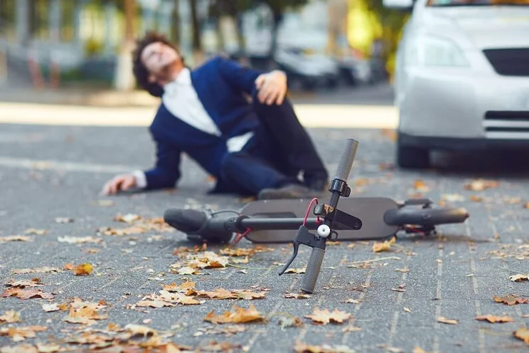 A man in a suit has fallen off of an electric scooter. There is a silver car to the right of the image, the e-scooter is in focus, the man in the suit is out of focus