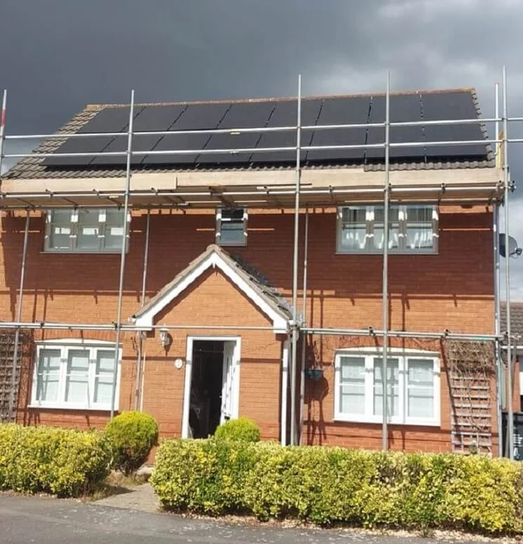 red brick detached house with scaffolding around it and solar panels on roof, dark clouds in background