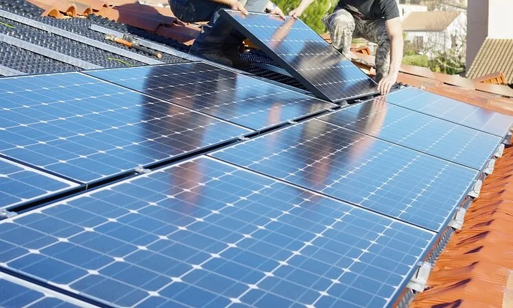 Two people installing rows of blue solar panels onto a roof with terracotta tiles