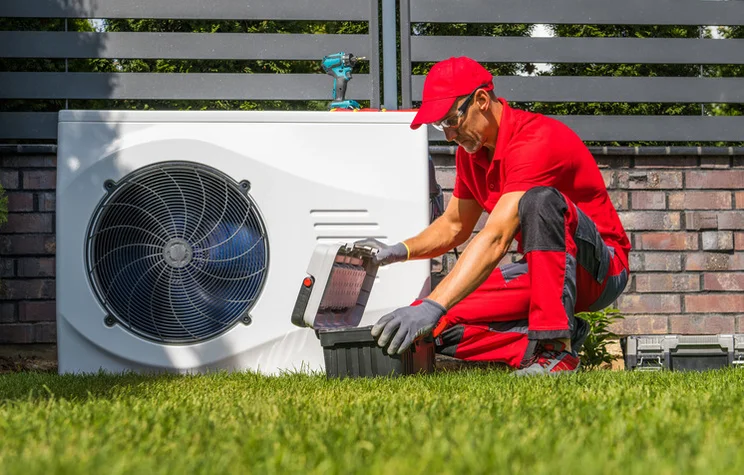 A workman squatting next to an outdoor heat pump, opening a tool box.
