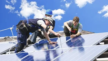 three people installing solar panels on a roof, against a blue sky