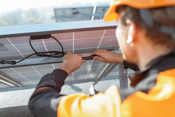 Installer in an orange hard hat and high vis jacket handling wires at the back of a solar panel