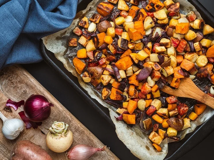 Roasted vegetables on a baking tray next to fresh unprepared vegetables on a chopping board