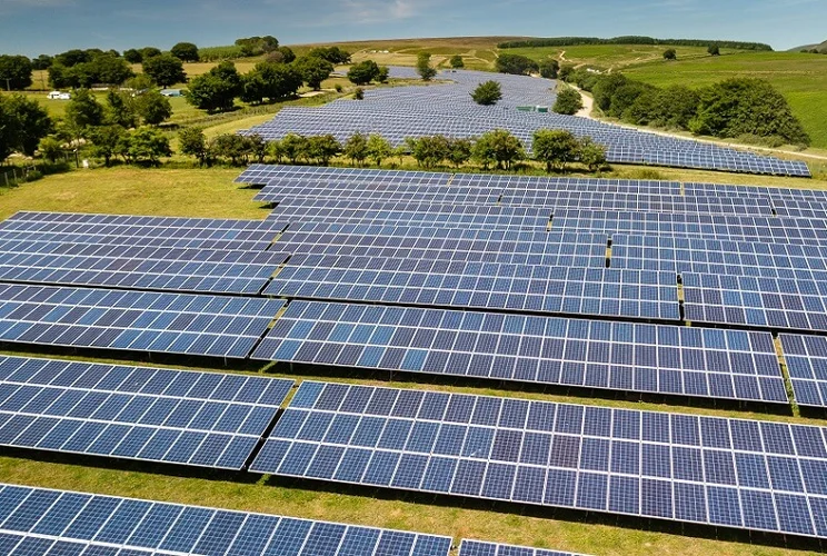 Aerial view of a solar farm operating in the Welsh countryside. There are many solar panels over a green field with a clear blue sky in the background.