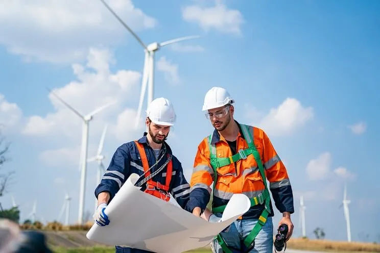 Two engineers in high vis jackets examining plans of a wind turbine farm with turbines in the background