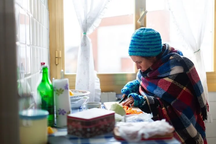 woman wrapped up in blanket and wool hat cutting carrots in kitchen