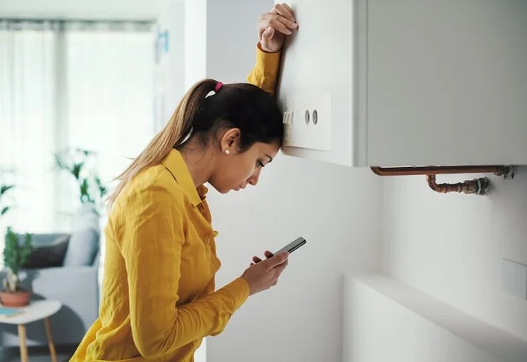 Woman leaning her head in frustration against a broken boiler as she looks at the phone in her hand