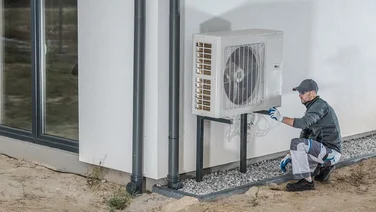 A man is installing an air source heat pump on the side of a modern home.