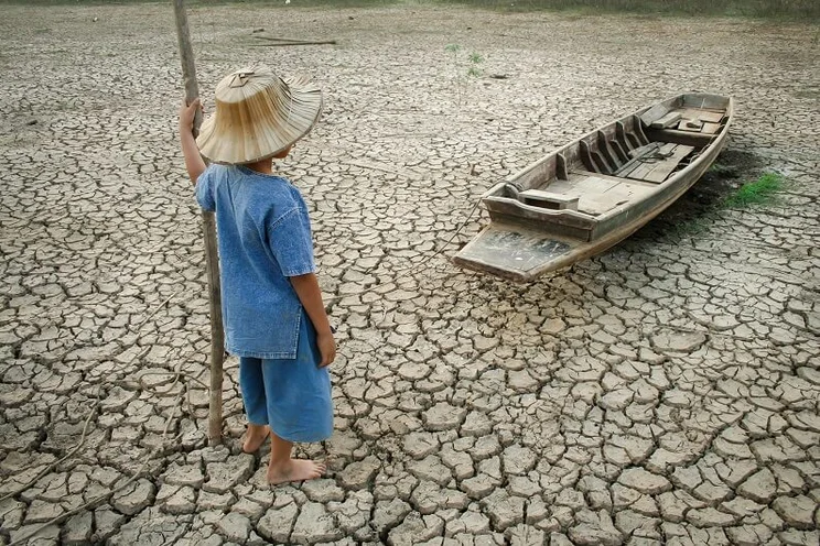 Boy staring at wooden boat resting on the cracked earth of a dried riverbed
