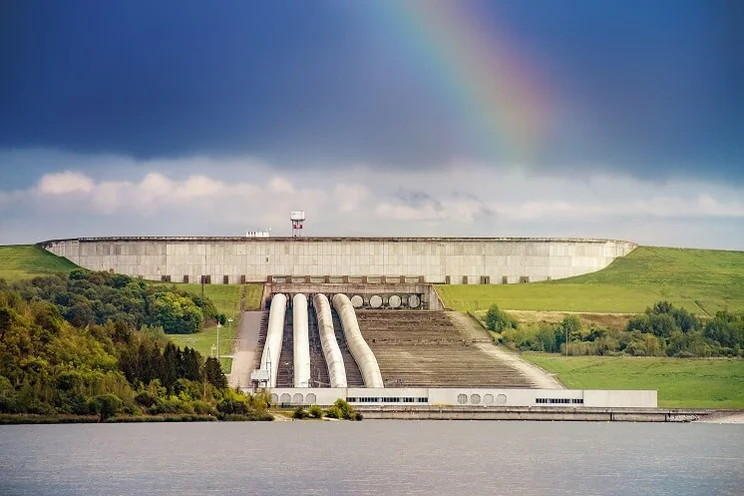 dam connected by pipes to hydroelectric station on hill above, dark sky and rainbow in background