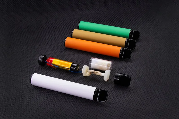 Five disposable vapes lined up, one has been taken apart to show the internal components including a lithium-ion battery