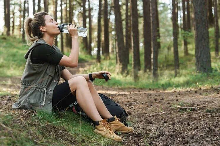 woman hiker sitting on ground in forest drinking out of reusable water bottle