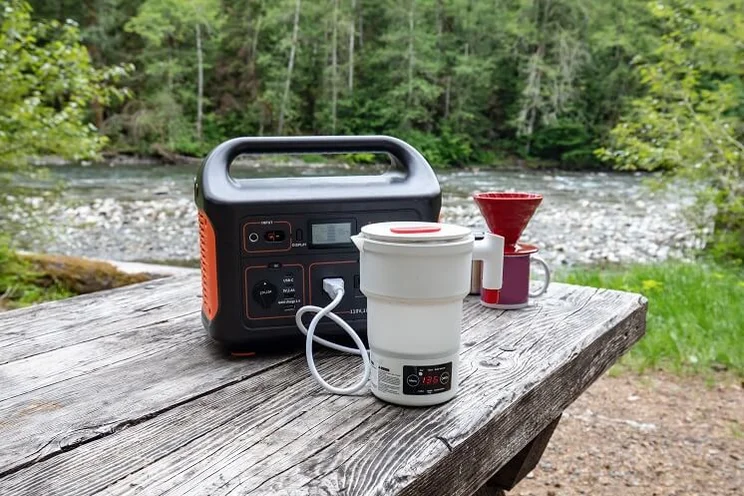 portable power generator charging kettle and small coffee machine on a wooden picnic table with trees and small lake in background