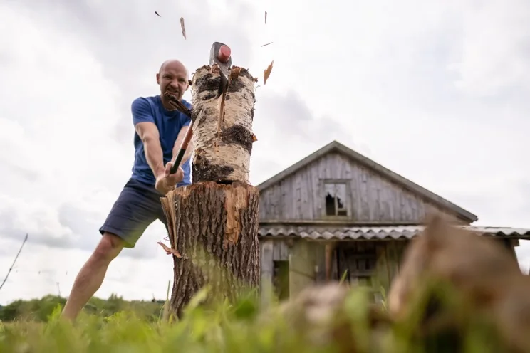 man chopping a log on a bigger log, in front of a barn and a white sky
