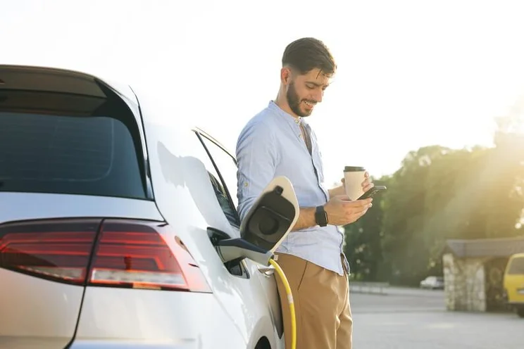 Man looking at phone smiling while electric car charges at a public charging point