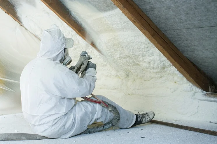 Installer dressed in white overalls sits down to install spray foam insulation in a hard-to-reach area