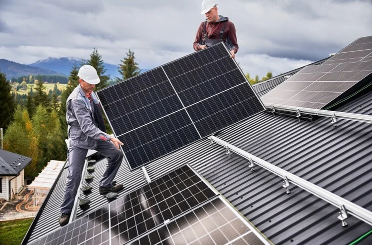 People installers carrying a solar panel on a roof with a cloudy sky in the background