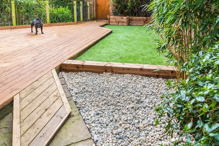 A garden with a small dog standing on wooden decking next to a patch of green artificial grass