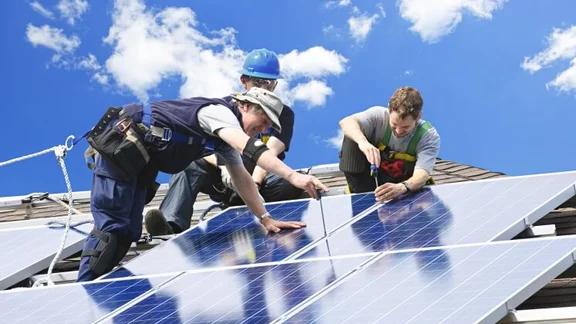 three people installing solar panels on a roof, against a blue sky