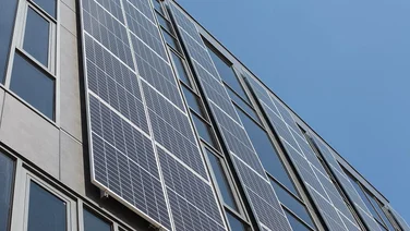 Lots of blue solar panels lined up vertically on the side of a building with windows on it