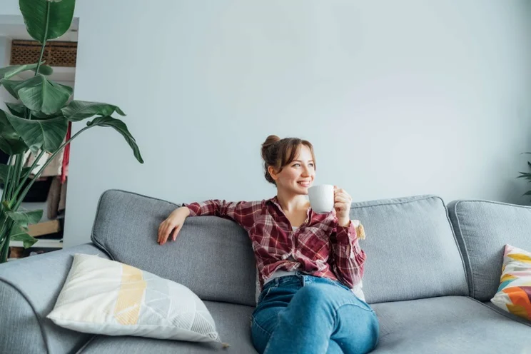 Young smiling woman sitting on sofa against a wall and looking away while holding a mug