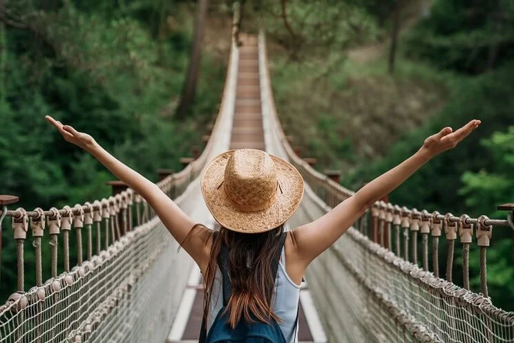 woman with arms outspread on suspended wooden bridge going through forest