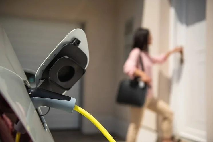 Electric car charging while woman wearing pink shirt and beige trousers enters her home