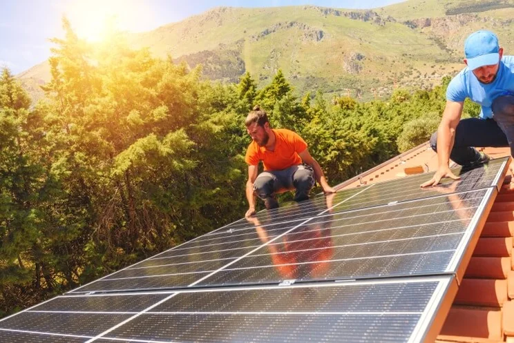 two people installing solar panels on a roof with trees and sunshine in the background