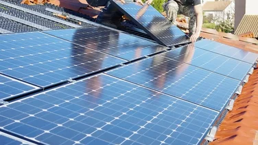 Two people installing rows of blue solar panels onto a roof with terracotta tiles