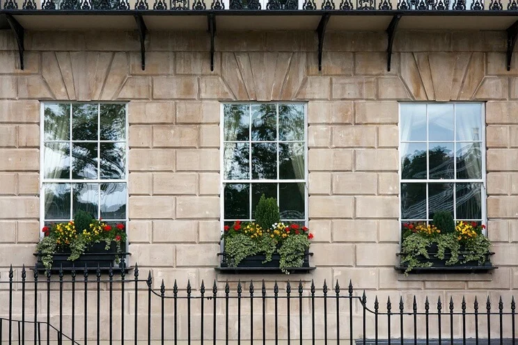 row of three white, rectangular sash windows with flower pots in front in brown stone building