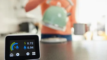 Close Up Of Smart Energy Meter In Kitchen Measuring Electricity And Gas Use With Woman Boiling Kettle